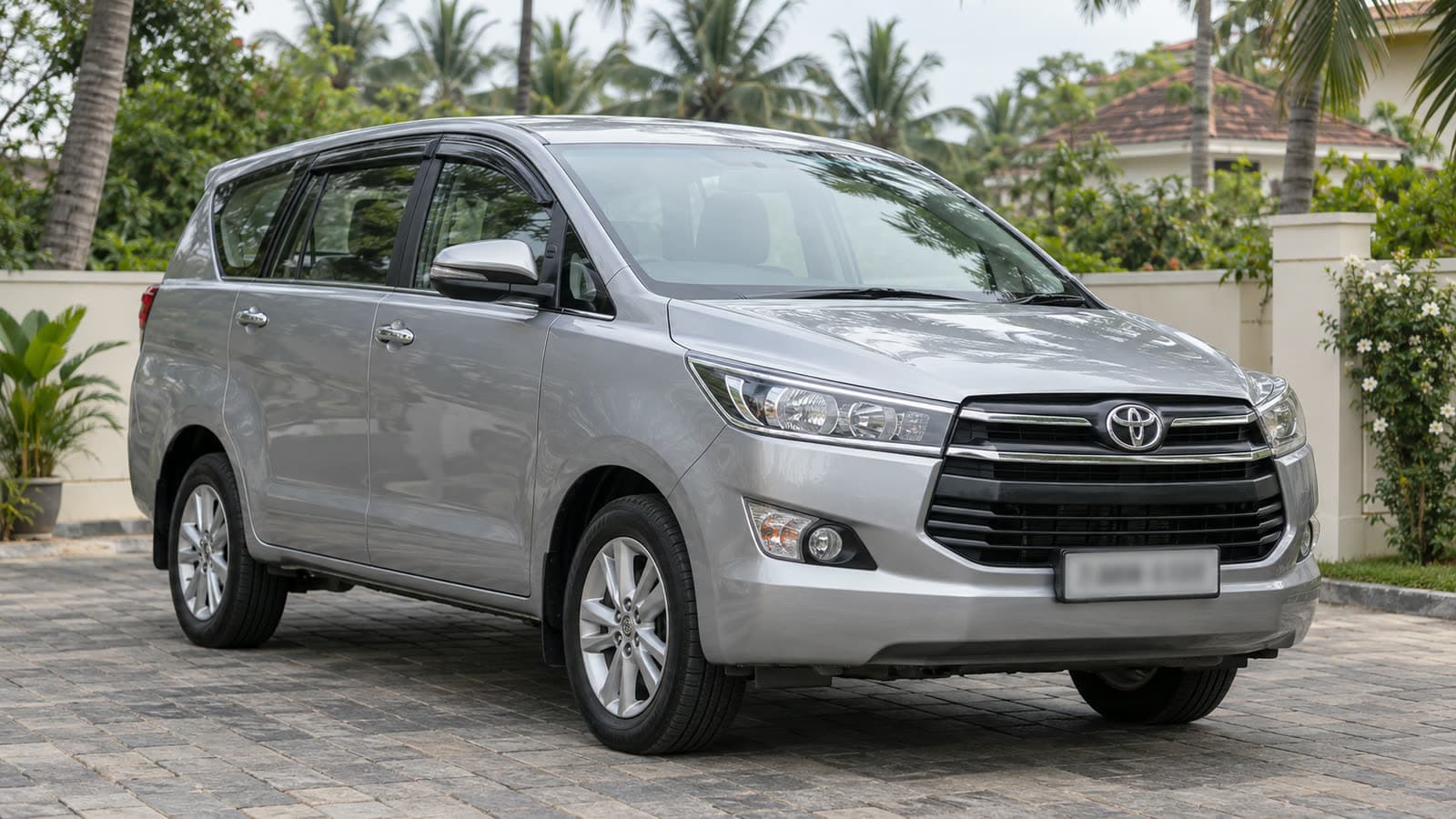 A silver Toyota Innova Crysta MPV parked outside a Kerala house in daylight, palm trees behind