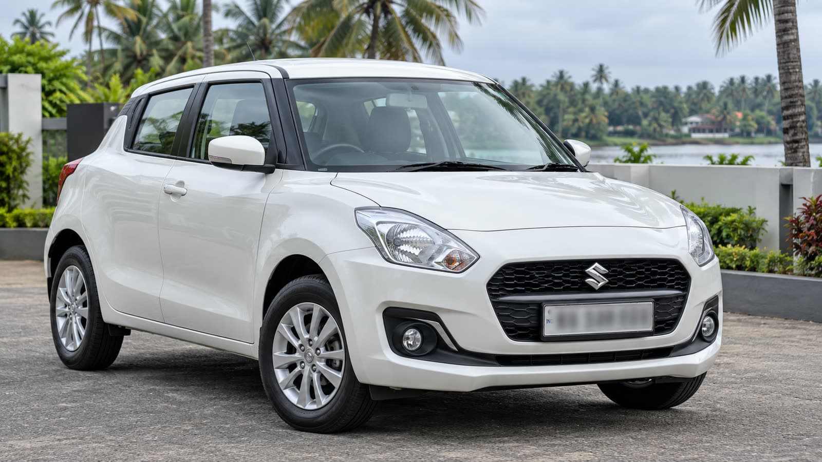 A white Maruti Swift hatchback parked on a Kerala driveway in daylight, with palm trees and a house behind it